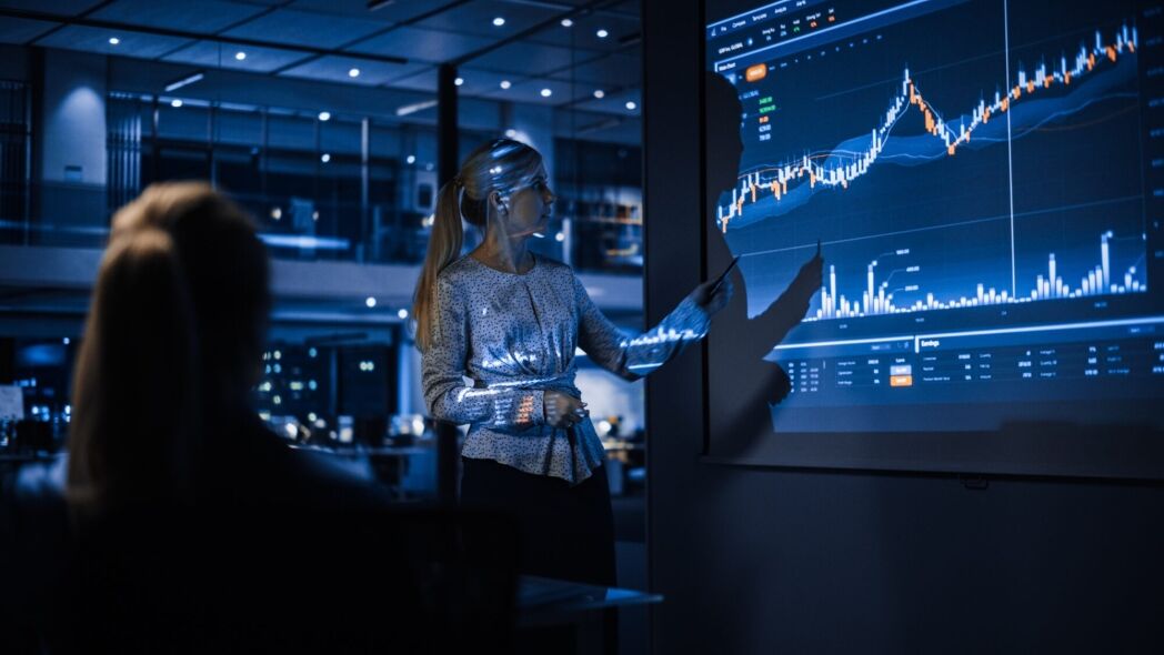 A woman in a business setting points to a financial chart on a large screen while another person is silhouetted in the