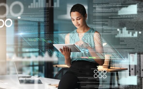 A woman in a teal top and black pants uses a tablet in an office environment with futuristic holographic data displays.