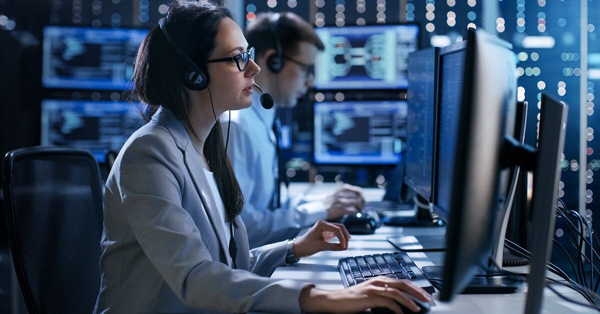 Two individuals wearing headsets and glasses work at computers in a dimly lit control room with multiple monitors