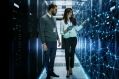 Two IT professionals, a man and a woman, stand in a server room, with the woman pointing at a holographic network