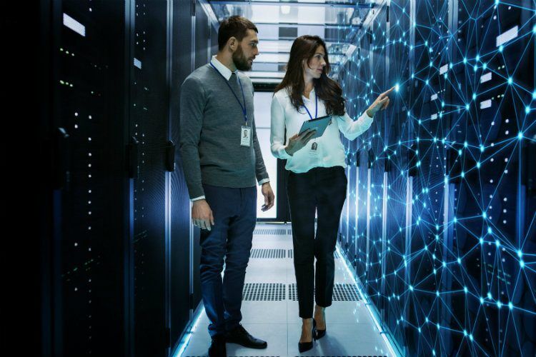 Two IT professionals, a man and a woman, stand in a server room, with the woman pointing at a holographic network
