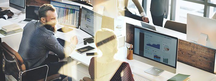 A man with a beard works at a computer in a modern office setting, with other colleagues visible in the background.