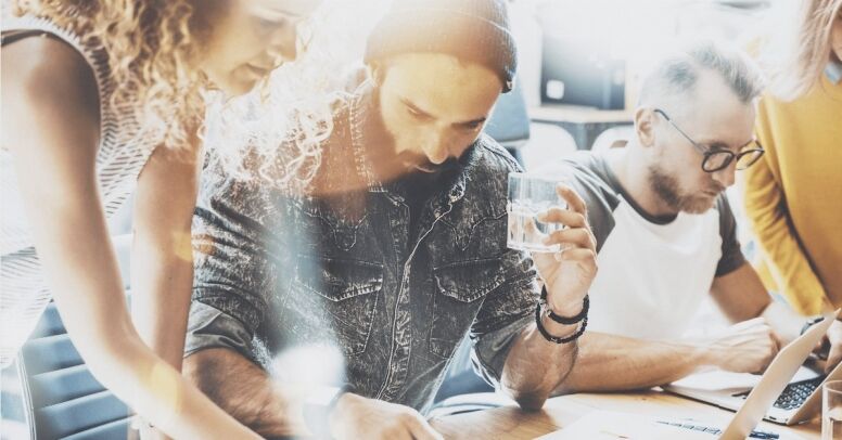 A group of colleagues collaborates around a table, focused on laptops and documents.