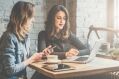 Two women are working on laptops and using their phones at a table in a cafe with a brick wall background.