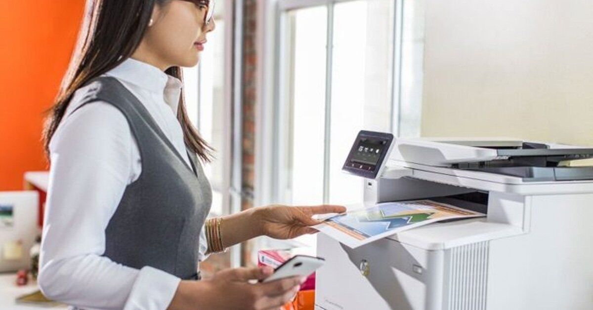 A woman in business attire uses a smartphone while interacting with a printer in an office setting.