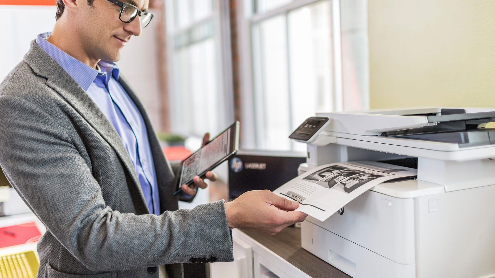 A man in a suit holds a tablet while retrieving a printed document from a printer.