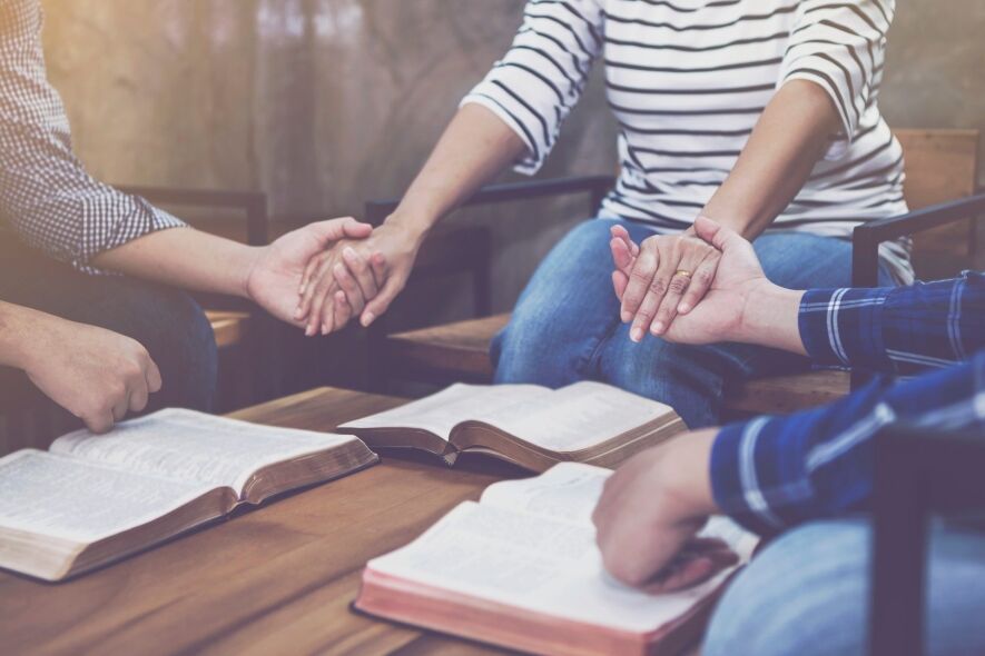 Three people hold hands over open Bibles on a wooden table, suggesting a prayer group or Bible study.
