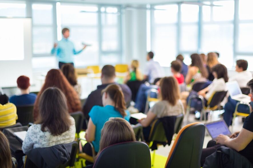 A presenter addresses an audience seated in a classroom or lecture hall.