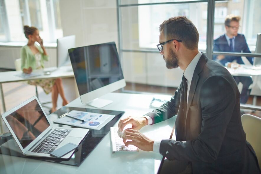 A man in a suit with glasses is typing on a keyboard in an office setting, with a laptop and charts on his desk.