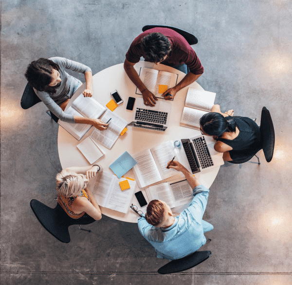 Four students are gathered around a circular table, engrossed in their studies with books, laptops, and notepads.