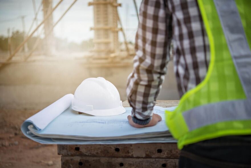 A construction worker in a safety vest reviews blueprints and a hard hat at a worksite.