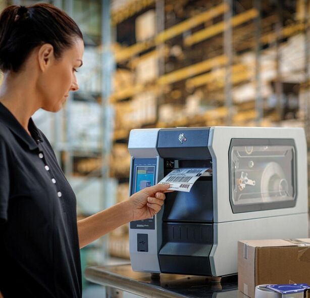 A woman in a warehouse is inserting a printed label into a Zebra label printer.