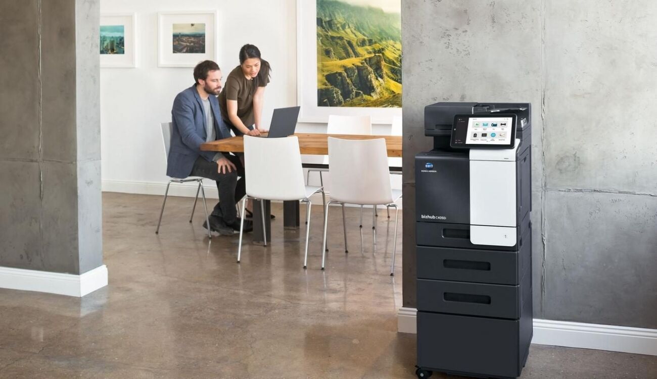 Two people collaborate on a laptop at a table next to a multifunction printer in a modern office space.