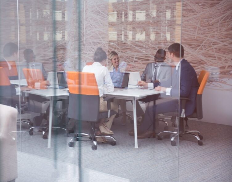 A diverse group of professionals are engaged in a meeting around a table, with laptops and papers suggesting a work
