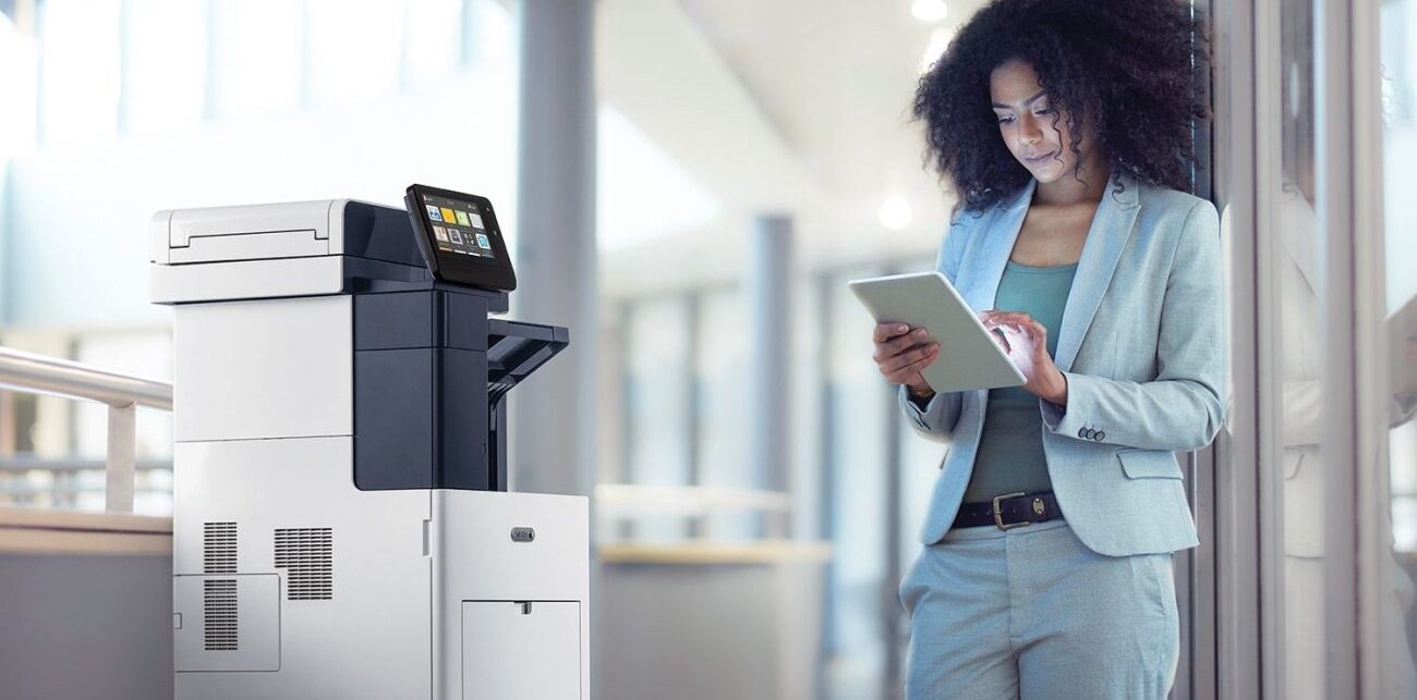 A woman in a light blue suit uses a tablet next to a large office printer.