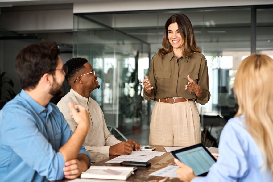 A female leader addresses a group of attentive colleagues during a business meeting in a modern office environment.