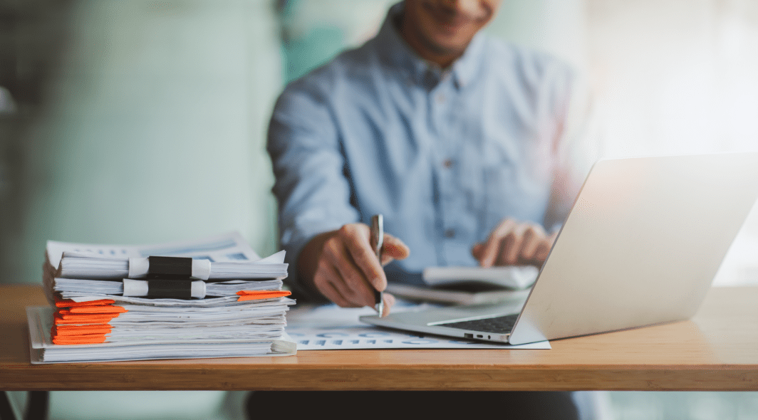 A person in a blue shirt is working at a desk with a stack of papers, a laptop, and a calculator.