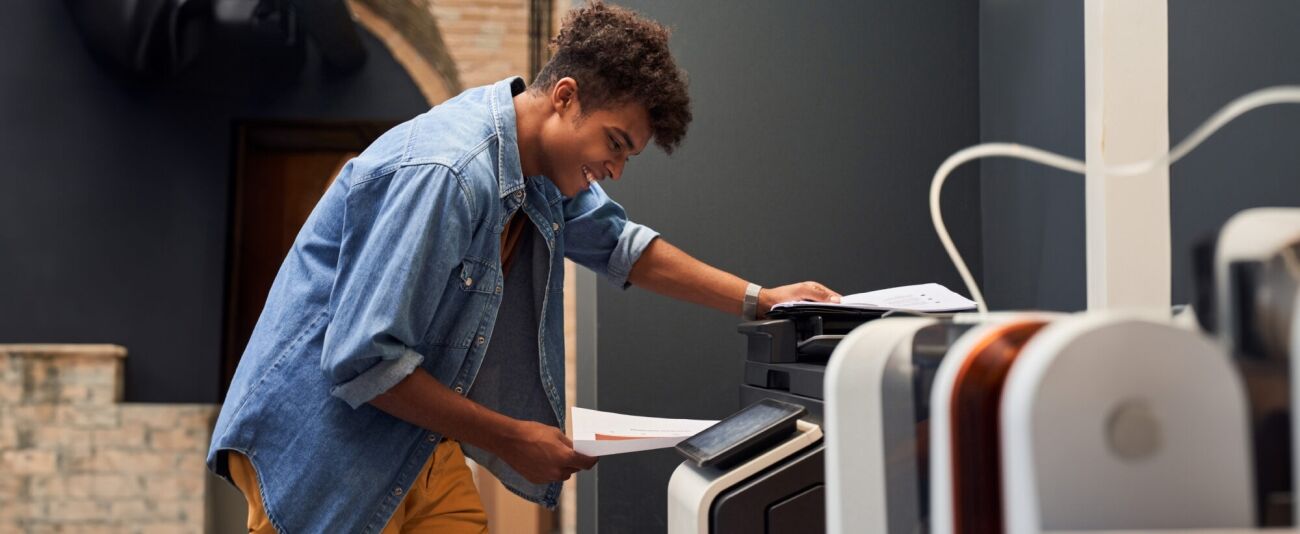 A young man smiles intently while operating a printer, holding documents in his hands.