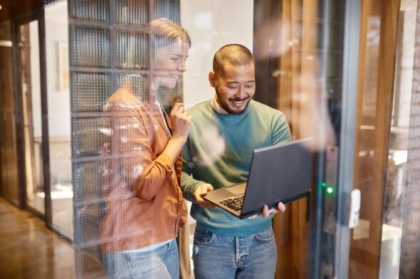A man and a woman smile while looking at a laptop together, captured through a reflective glass wall in a contemporary