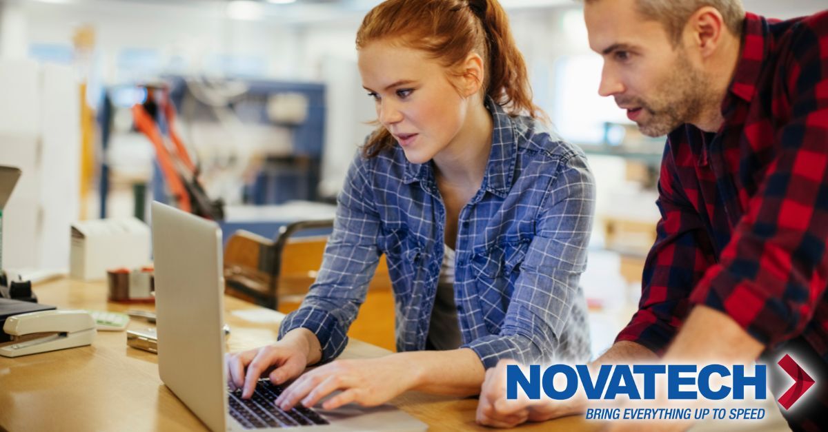A young woman with red hair and a man in a plaid shirt collaborate on a laptop in a workshop setting.
