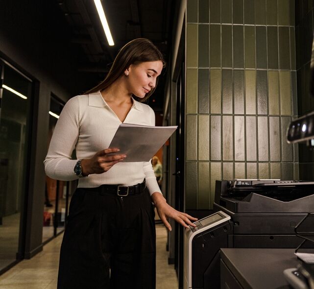 A young woman operates a printer in an office setting while holding a stack of papers.