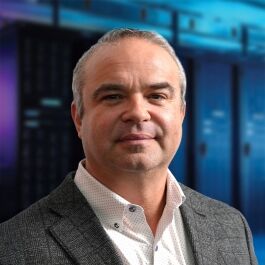 A man in a blazer and patterned shirt smiles in