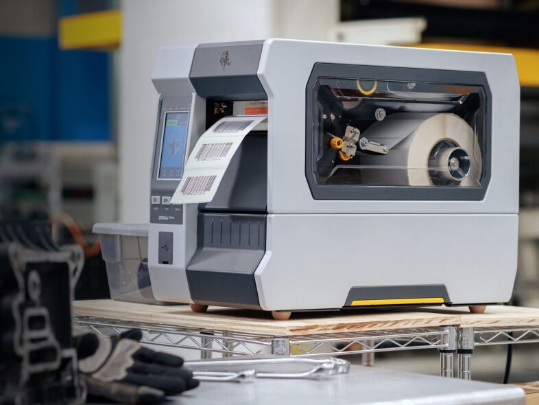 A Zebra ZT610 industrial label printer is shown on a shelf, printing a continuous roll of labels.