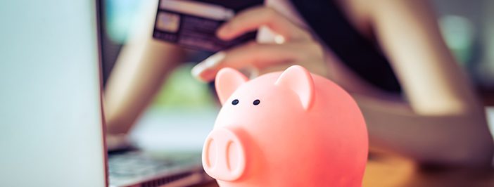 A person uses a credit card while a piggy bank sits in the foreground, suggesting online shopping or financial