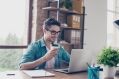 A man in glasses and a teal shirt is working on a laptop and drinking from a coffee cup at a wooden desk.