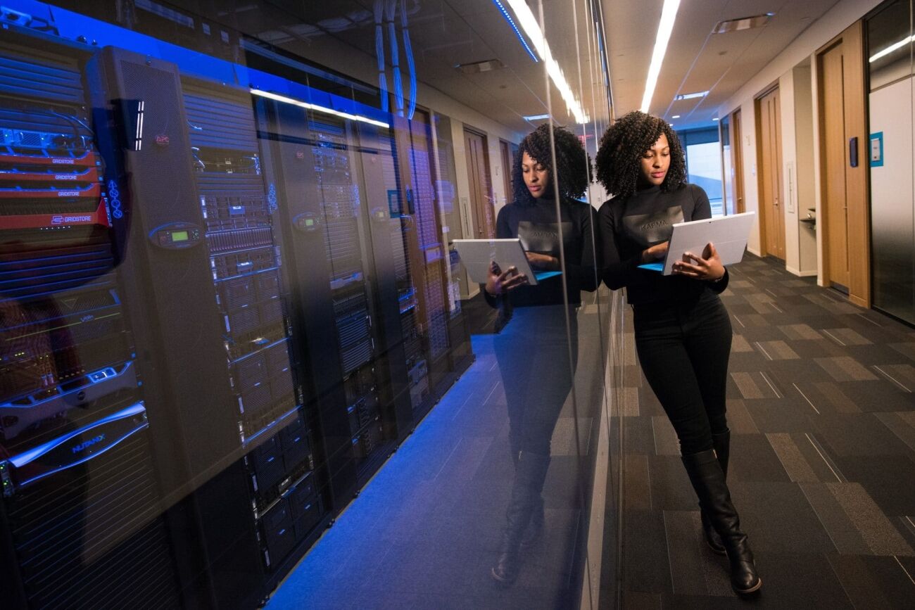 A woman in black attire stands in a server room, looking at a tablet.
