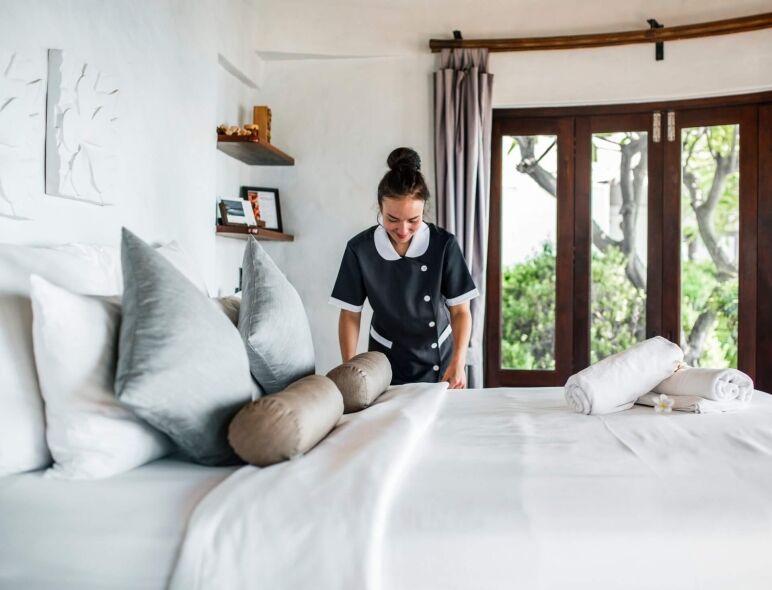 A smiling housekeeper in uniform meticulously tidies a freshly made bed in a bright, modern hotel room.