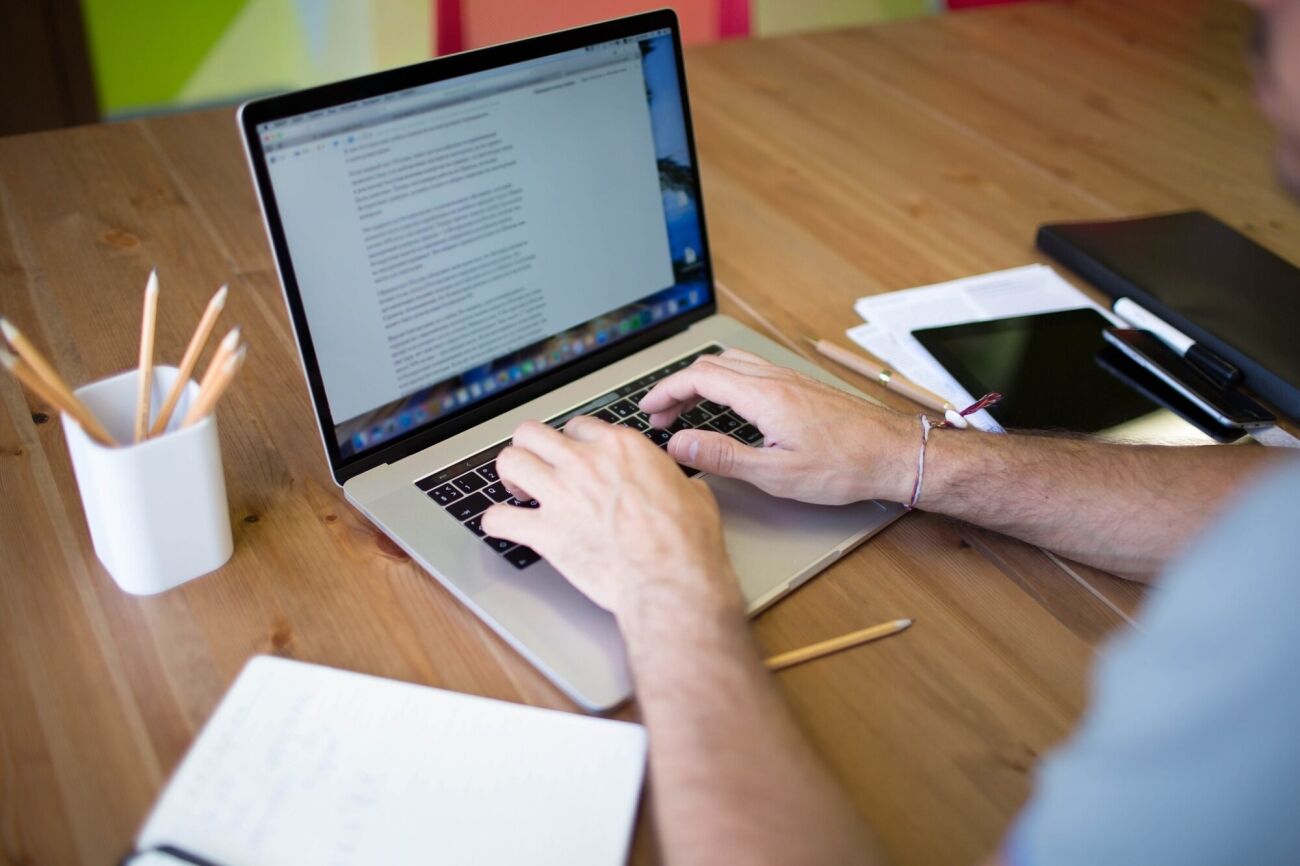 A person's hands are typing on a laptop on a wooden desk, surrounded by office supplies.