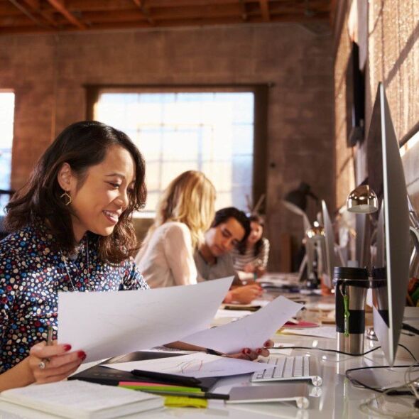 A woman in a patterned shirt smiles as she reviews documents at her desk in a modern office with colleagues working in