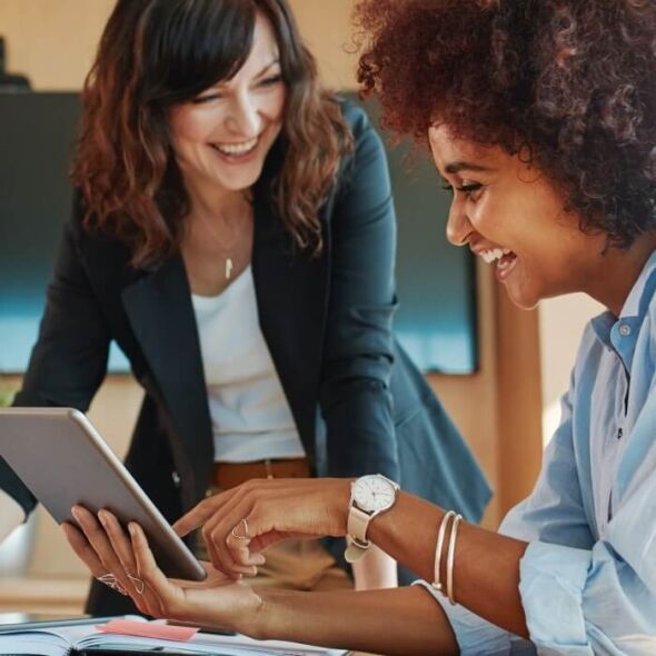 Two women in business attire laugh while looking at a tablet.