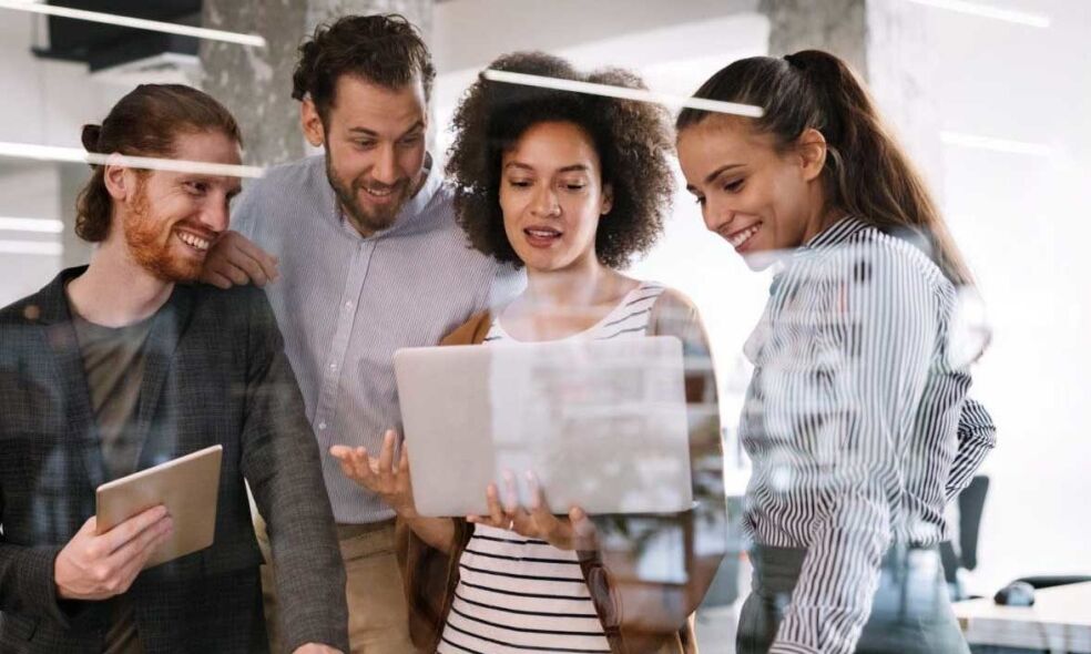 Four colleagues are gathered around a laptop, engaged in a discussion in a modern office setting.