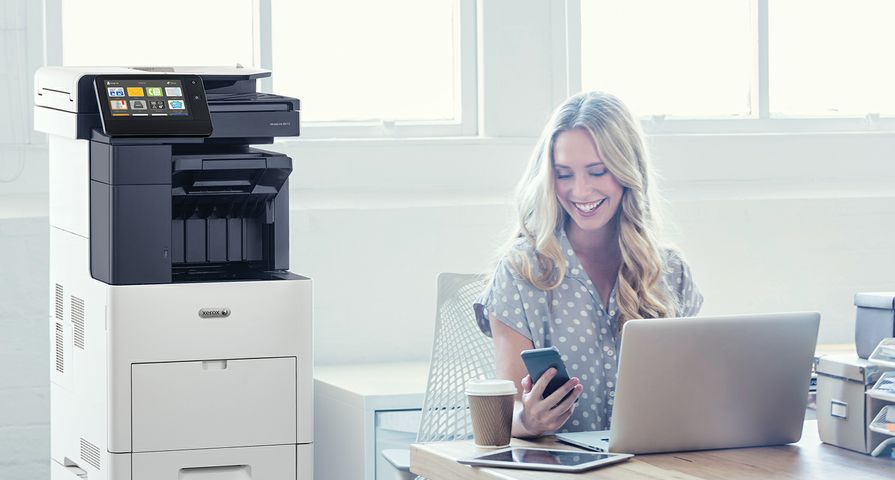 A woman smiles while looking at her phone next to a Xerox printer and a laptop.
