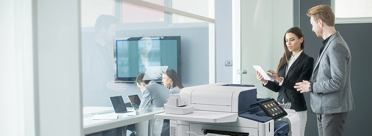 Two business professionals, a woman and a man, are looking at a tablet and a printer in a modern office setting.