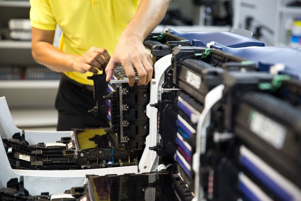 A technician in a yellow shirt services the intricate internal components of a large commercial printer.