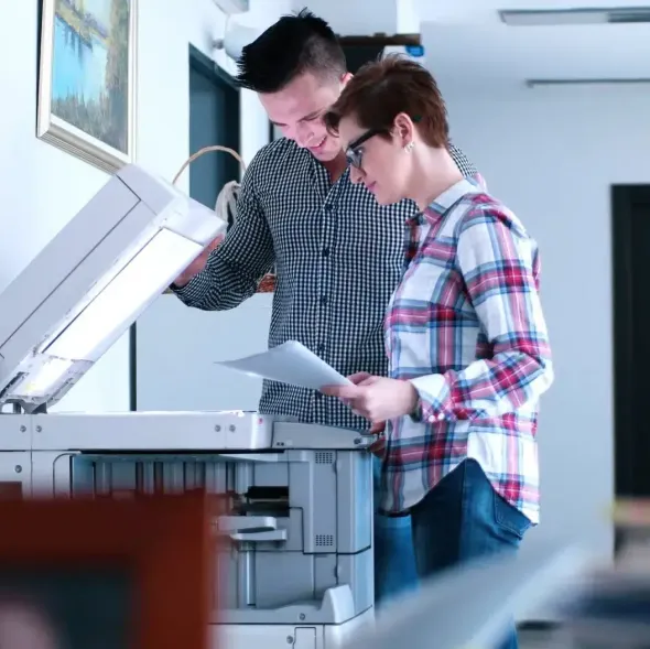A man and a woman are collaborating to use a large office photocopier, with the man lifting the lid and the woman