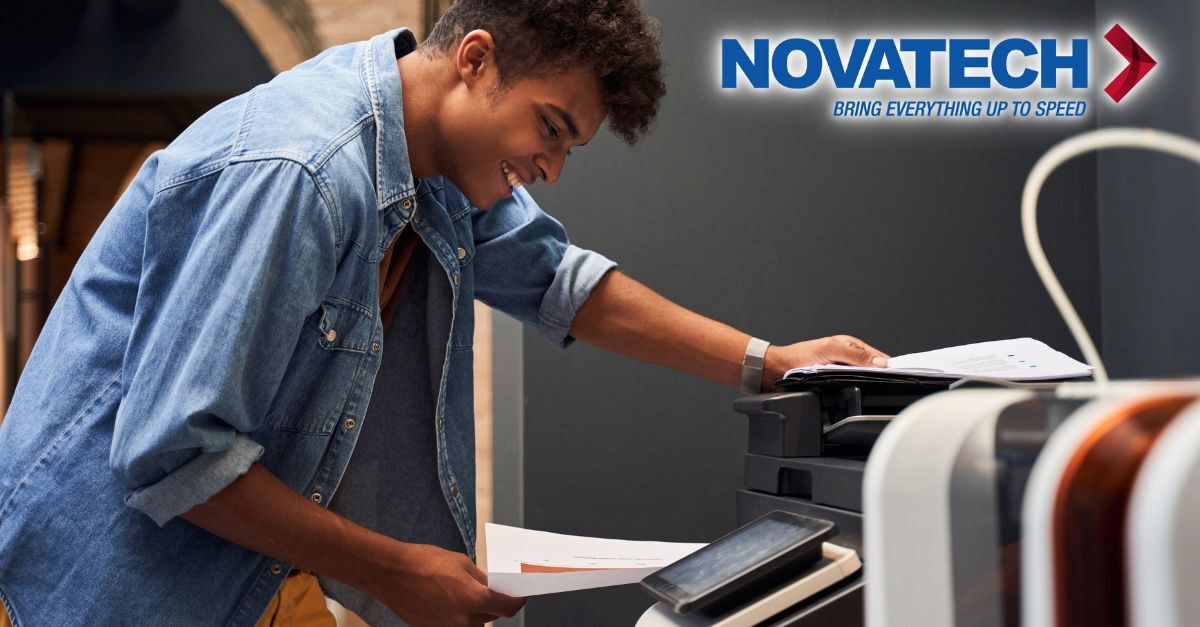 A young man in a denim jacket smiles as he places papers into a printer, with the Novatech logo visible in the
