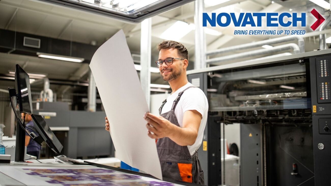 A smiling man in overalls holds a large sheet of paper while standing in a printing facility next to a large printing