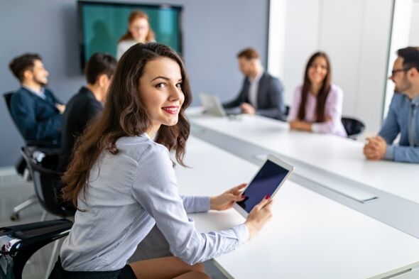 A young woman smiles at the camera while holding a tablet in a business meeting with colleagues in the background.