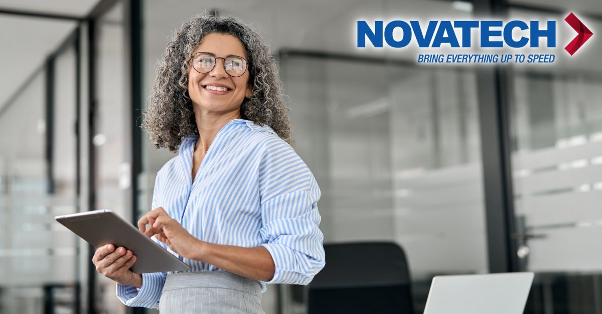 A smiling woman with curly gray hair and glasses holds a tablet in a modern office setting, with the Novatech logo