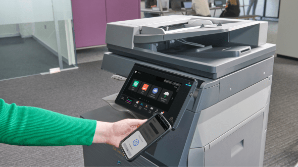 A person's arm in a green sweater holds a smartphone near the touchscreen of a large office copier.