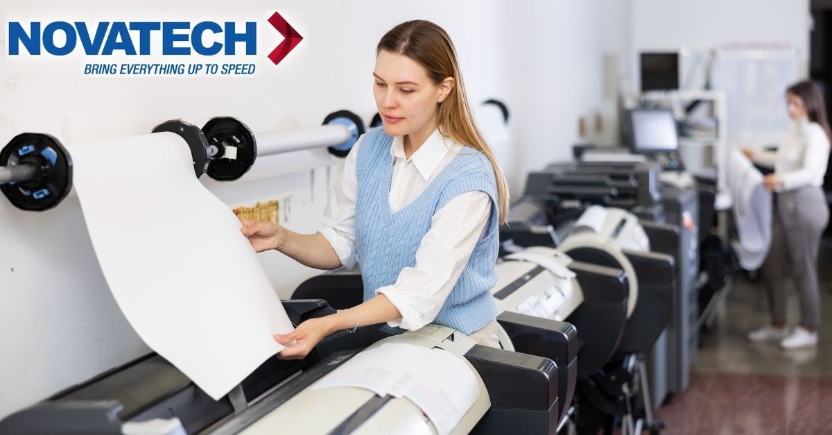 A woman in a blue vest and white shirt operates a large format printer, with a roll of paper feeding into the machine.