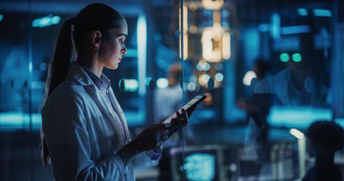A scientist in a lab coat uses a tablet in a dimly lit, futuristic laboratory environment.