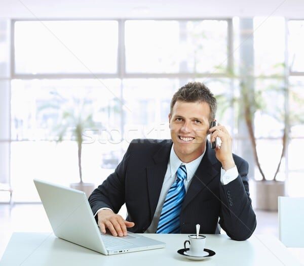 A smiling businessman in a suit talks on his cell phone while sitting at a desk with a laptop and coffee.