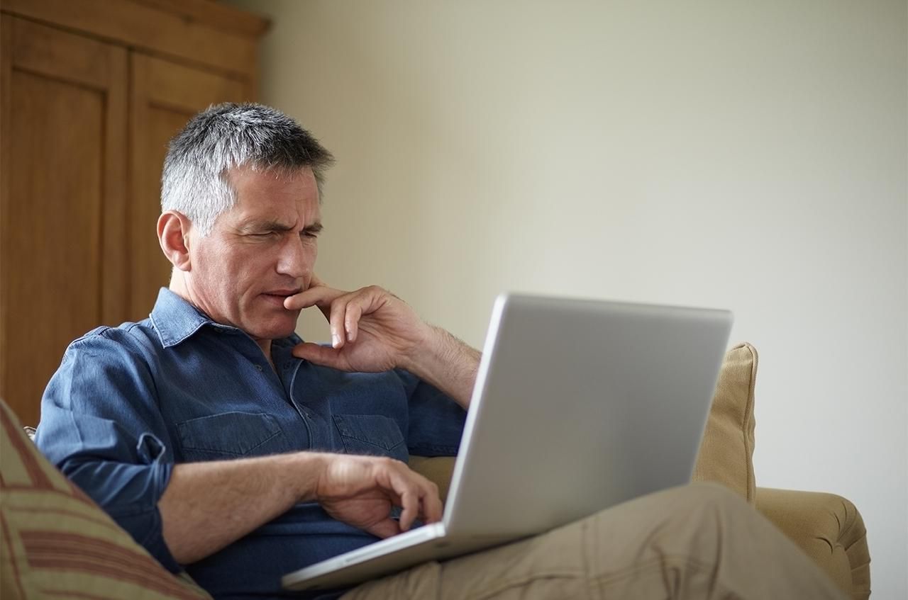 A middle-aged man with graying hair sits on a couch, looking intently at a laptop with his hand on his chin.