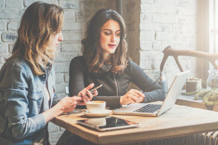 Two women are working on laptops and using their phones at a table in a cafe with a brick wall background.