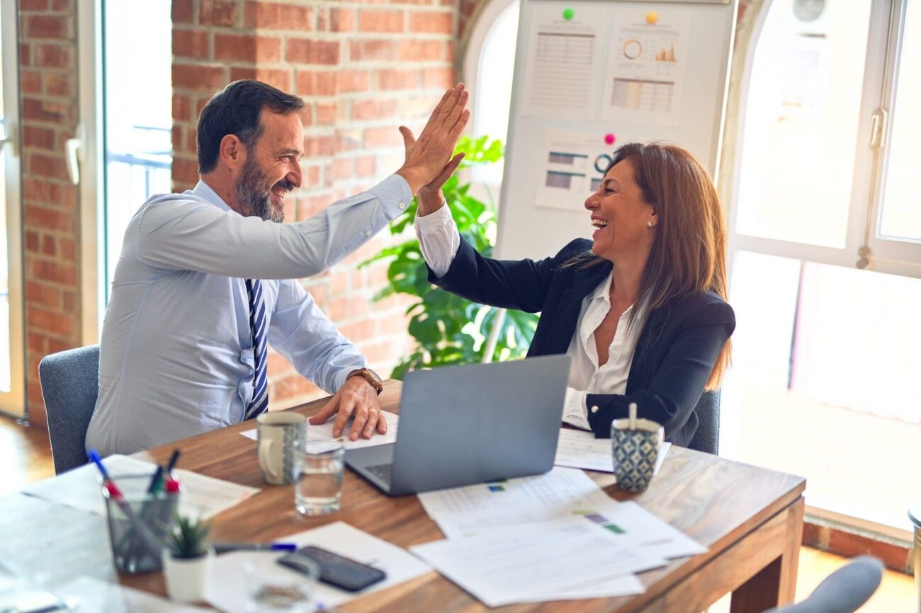 A man and a woman high-five each other while sitting at a desk in an office.